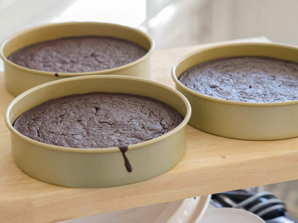 The BEST Vegan Chocolate Cake Recipe On The Internet. 19 Three round cake pans filled with freshly baked chocolate cakes rest on a wooden surface, with one cake showing a small drip of batter down the side of the pan. Bright natural light enters from the background.