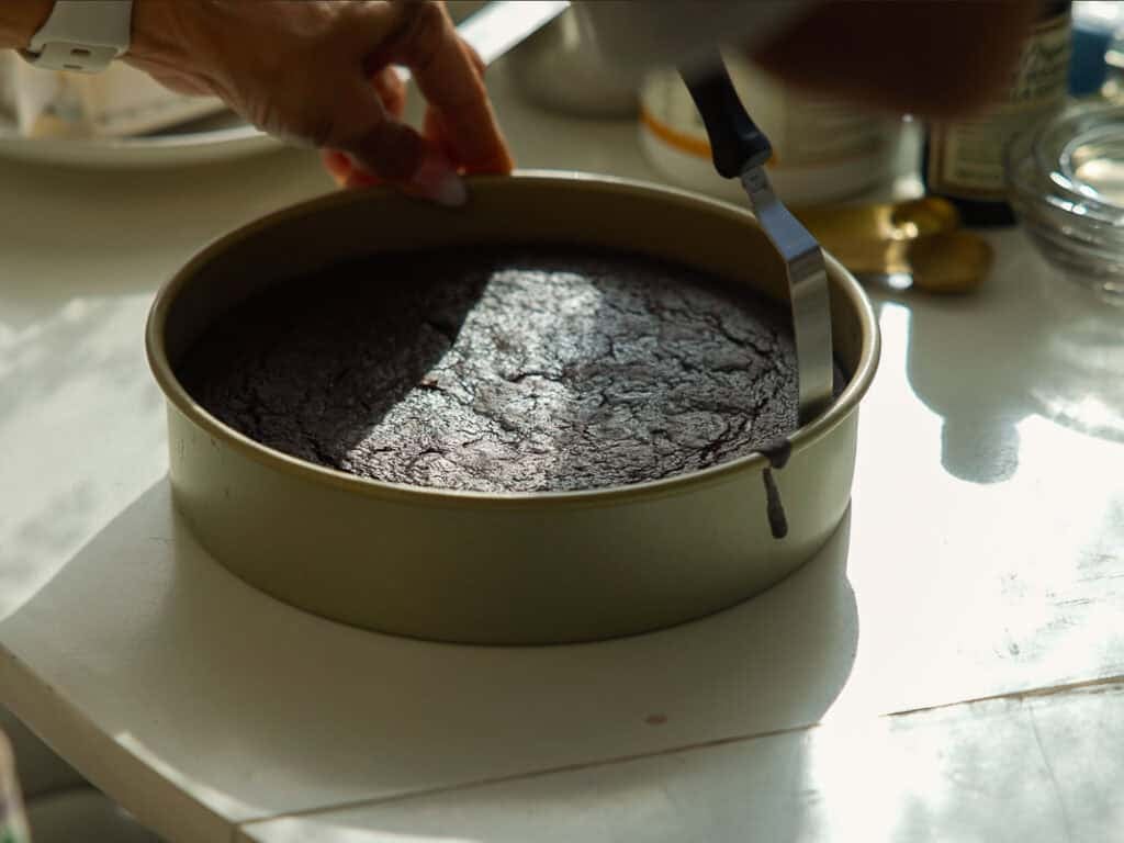 The BEST Vegan Chocolate Cake Recipe On The Internet. 20 A person uses a spatula to loosen the edges of a round chocolate cake in a baking pan on a sunlit kitchen counter.