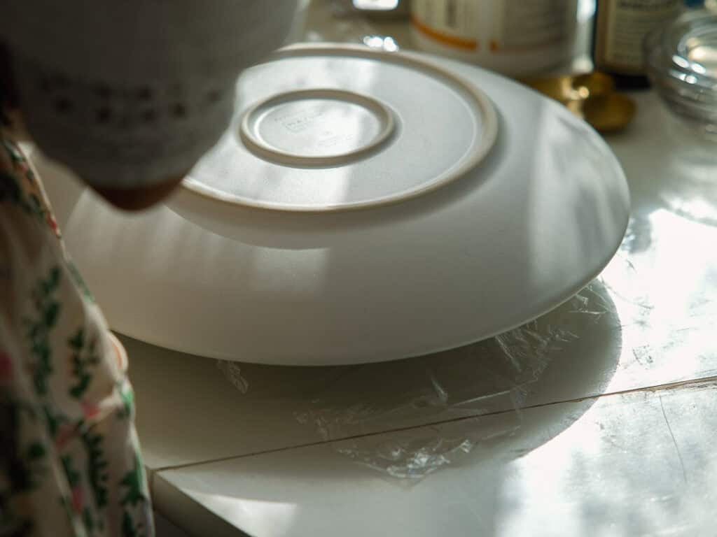 The BEST Vegan Chocolate Cake Recipe On The Internet. 21 A person covers a bowl with a large white plate on a sunlit kitchen counter, surrounded by food containers and utensils.