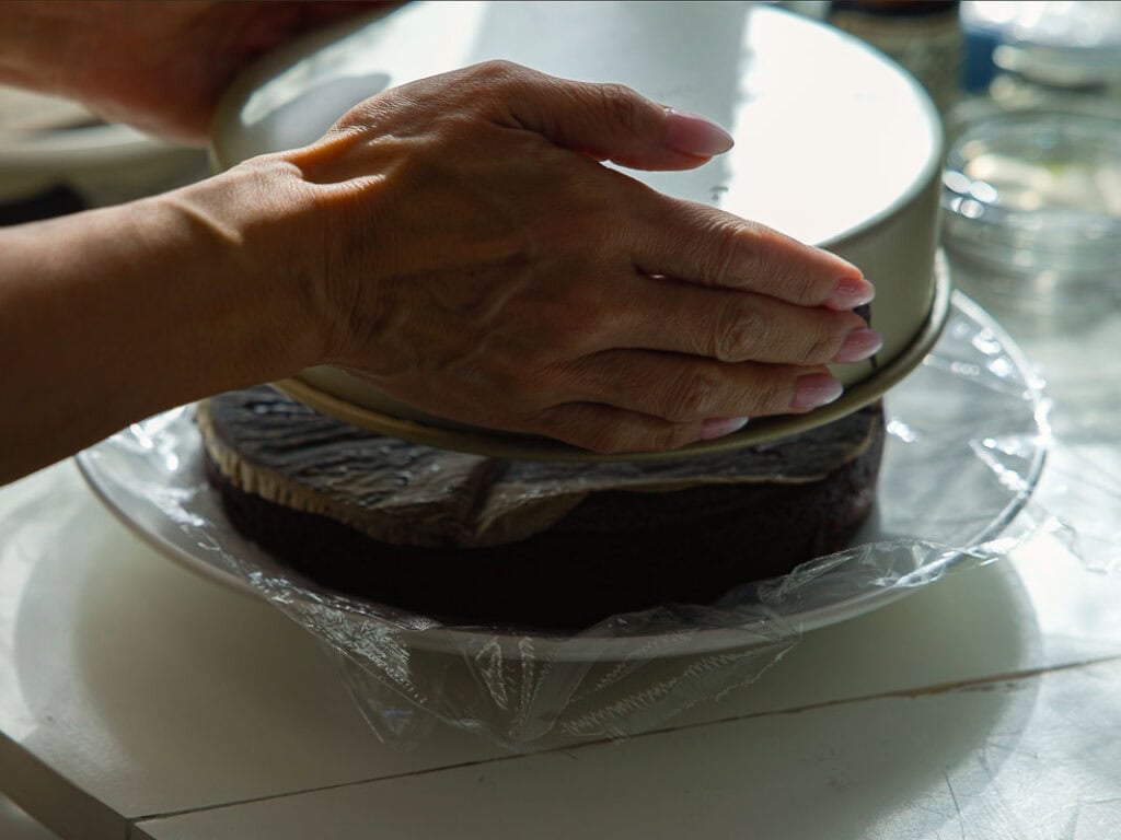 The BEST Vegan Chocolate Cake Recipe On The Internet. 22 A person removes a round cake pan placed over a chocolate cake on a plate, with plastic wrap partially covering the cake. Sunlight highlights their hands and the baking process.