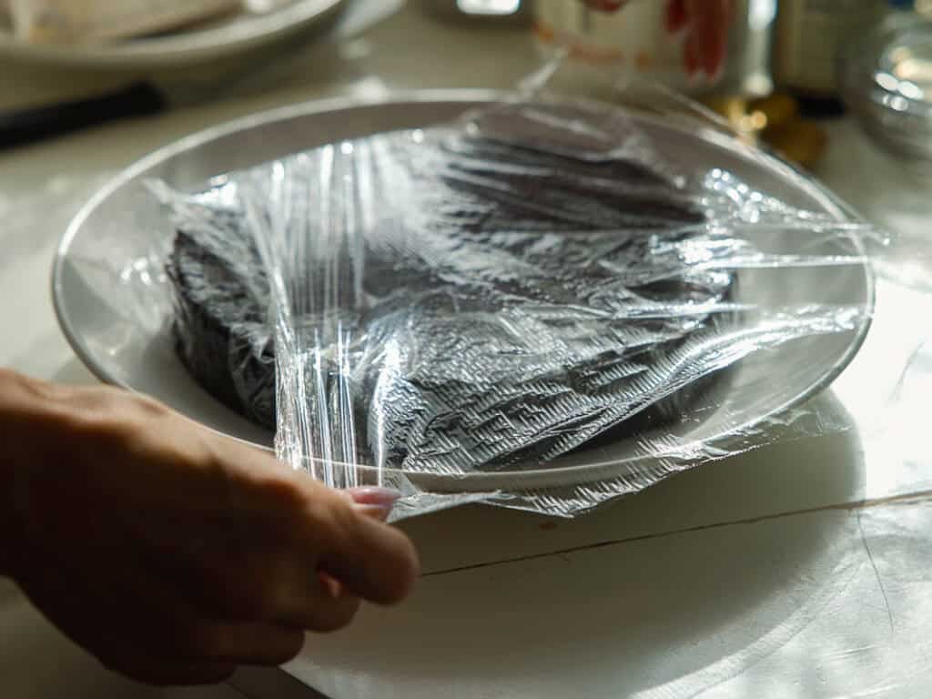 The BEST Vegan Chocolate Cake Recipe On The Internet. 24 A person’s hands are covering or uncovering a plate of dark-colored cake or brownies with clear plastic wrap on a white table.
