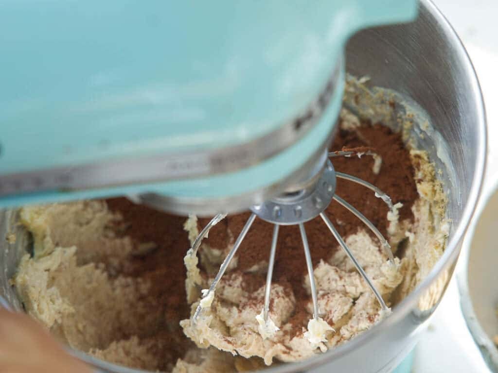 The BEST Vegan Chocolate Cake Recipe On The Internet. 25 A close-up of an electric stand mixer with a whisk attachment mixing vegan butter and cocoa powder in a stainless steel bowl. The mixer is light blue.