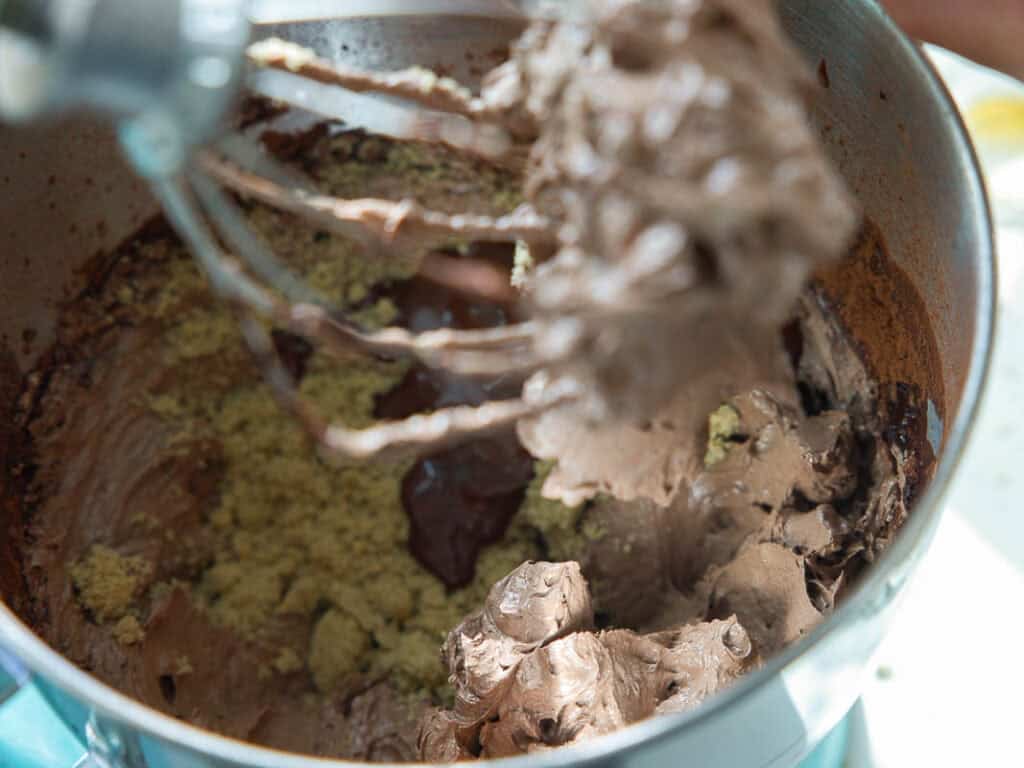 The BEST Vegan Chocolate Cake Recipe On The Internet. 26 A close-up of a stand mixer bowl with chocolate buttercream frosting and brown sugar being mixed, with some batter on the whisk attachment above the bowl. Bright daylight illuminates the scene.