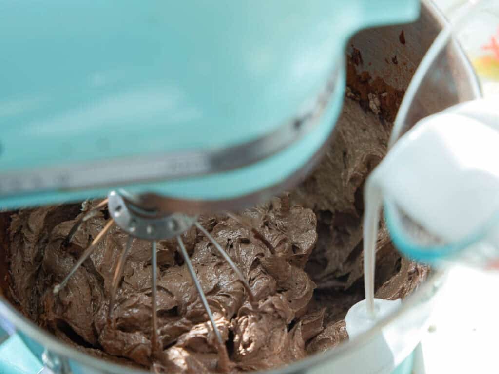 The BEST Vegan Chocolate Cake Recipe On The Internet. 27 A close-up of a light blue stand mixer whipping chocolate buttercream frosting with a metal whisk attachment, viewed from above. The mixture appears thick and creamy inside the mixing bowl.