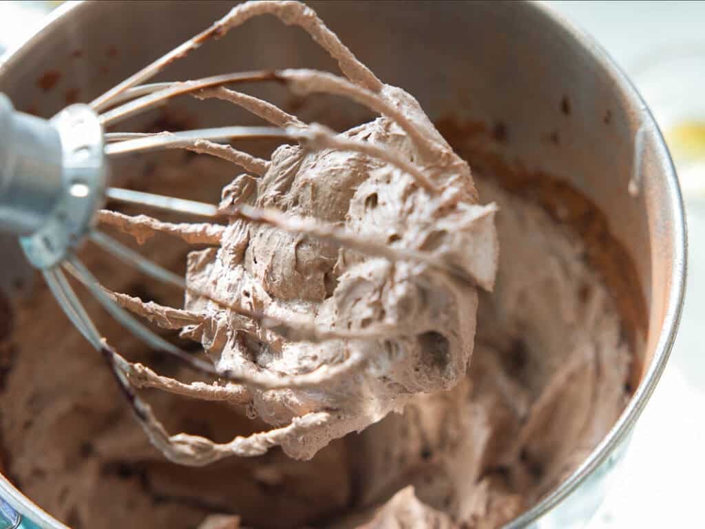 The BEST Vegan Chocolate Cake Recipe On The Internet. 28 A close-up of a metal whisk attachment covered in fluffy chocolate frosting, held above a mixing bowl filled with more chocolate frosting.