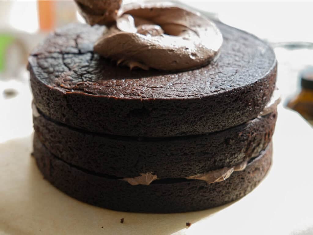 The BEST Vegan Chocolate Cake Recipe On The Internet. 32 Three layers of rich chocolate cake stacked with chocolate frosting between them, with a large dollop of frosting being spread on top. The cake sits on a light-colored surface.