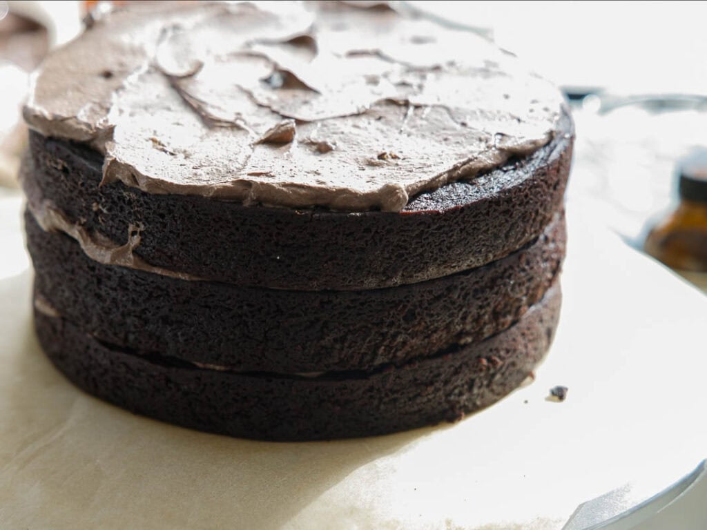 The BEST Vegan Chocolate Cake Recipe On The Internet. 33 A close-up of a three-layer chocolate cake with chocolate frosting spread on top, sitting on parchment paper. The cake layers are dark and moist, and there is no decoration or toppings.