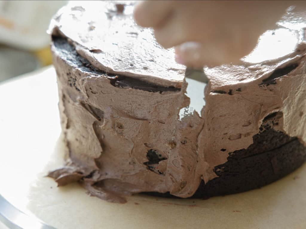 The BEST Vegan Chocolate Cake Recipe On The Internet. 34 A close-up of a hand spreading chocolate frosting on a round chocolate cake, partially covered with thick, creamy icing on a parchment-lined surface.