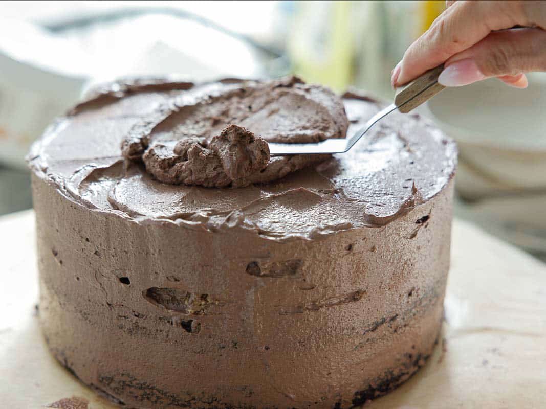 The BEST Vegan Chocolate Cake Recipe On The Internet. 35 A hand uses a spatula to spread chocolate frosting on a round, partially decorated chocolate cake.
