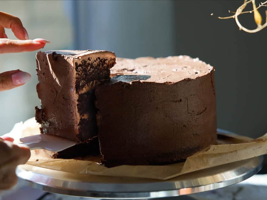 The BEST Vegan Chocolate Cake Recipe On The Internet. 3 A close-up of a hand lifting a thick slice from a round vegan chocolate cake with rich chocolate frosting, sitting on parchment paper and a metal tray in natural sunlight.