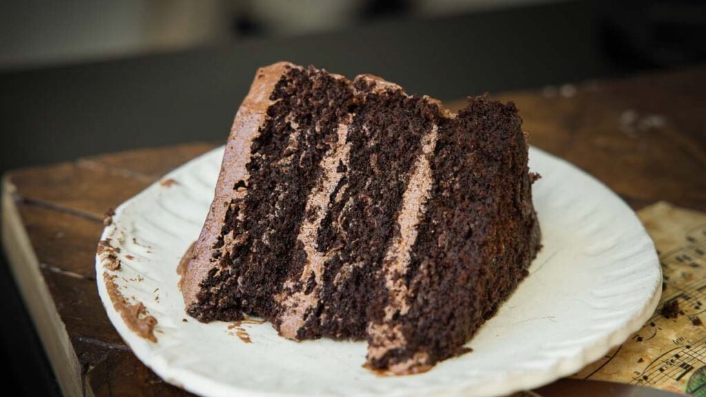 A slice of rich, three-layer vegan chocolate cake with chocolate frosting sits on a white plate atop a rustic wooden surface.