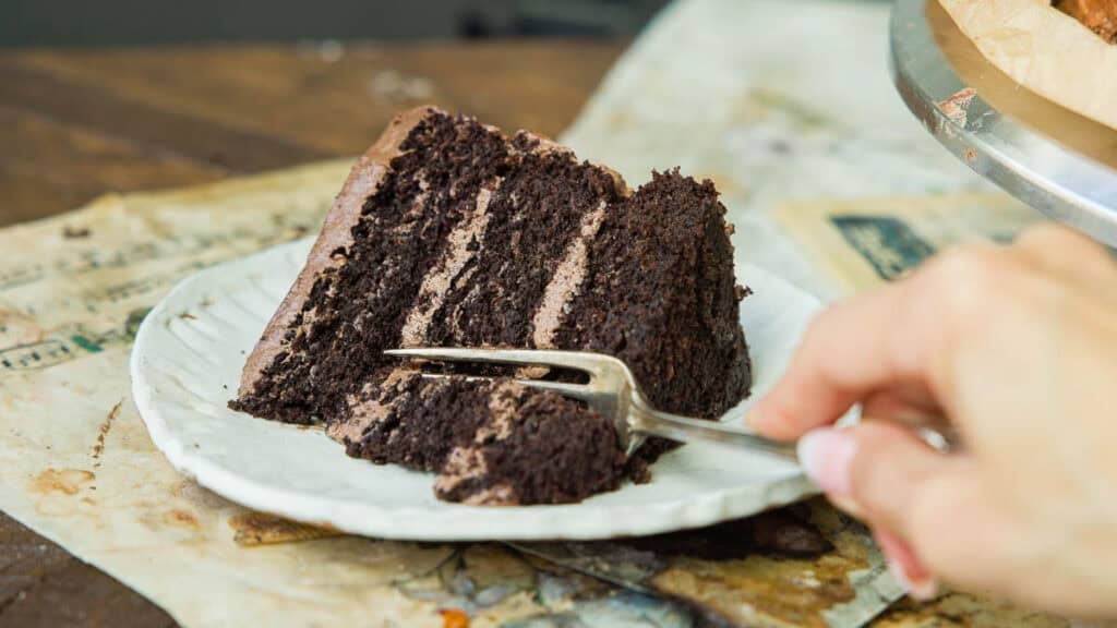The BEST Vegan Chocolate Cake Recipe On The Internet. 12 A hand holding a fork is about to take a bite from a slice of layered vegan chocolate cake on a white plate, set on top of old papers on a rustic wooden table.