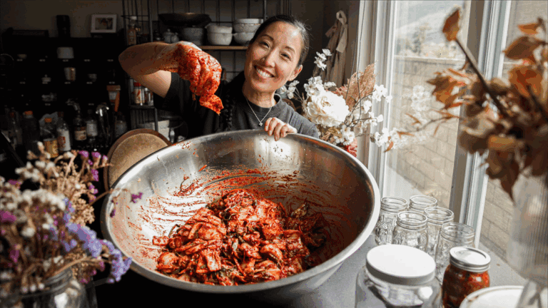 Recipes 11 Joanne Lee Molinaro holds up a piece of kimchi over a large metal bowl filled with more kimchi, surrounded by jars, flowers, and kitchen items—perfect inspiration for new recipes using kimchi—on a table by a window.