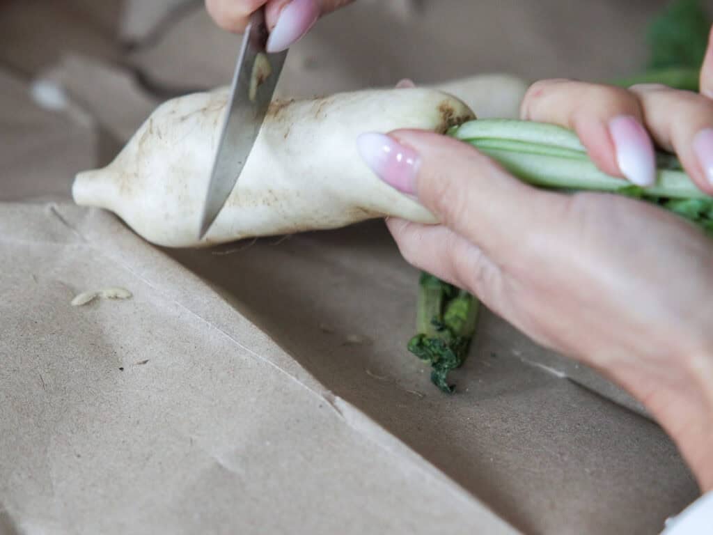 Vegan Ponytail Kimchi aka Chonggak-Kimchi Recipe. 16 A person with manicured nails holds a white radish and uses a knife to peel its skin over brown paper, preparing ingredients for a homemade Vegan Kimchi recipe.