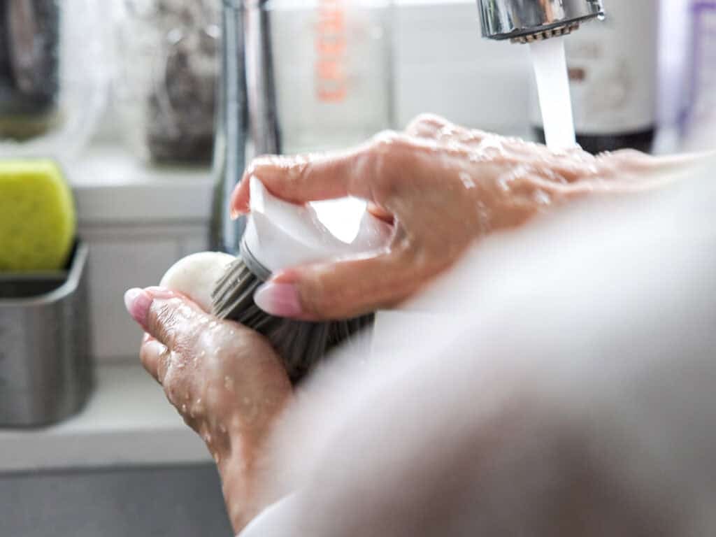Vegan Ponytail Kimchi aka Chonggak-Kimchi Recipe. 18 A person uses a scrub brush to clean a white vegetable, preparing it for a Vegan Kimchi recipe, under running water at a kitchen sink, with water droplets visible on their hands and the brush.
