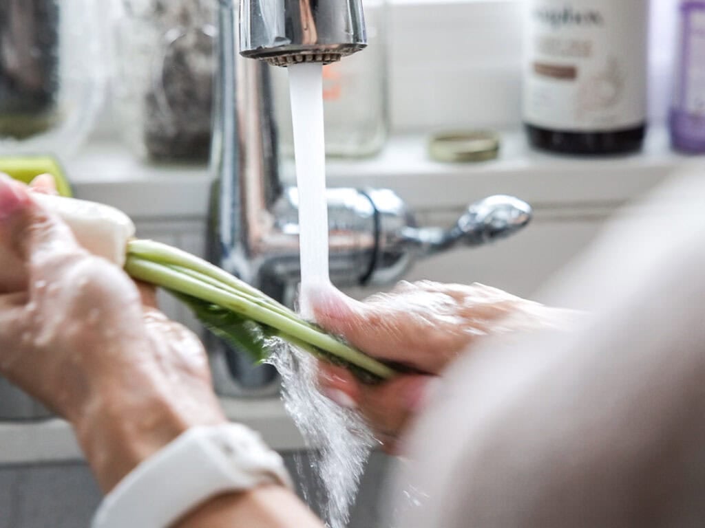 Vegan Ponytail Kimchi aka Chonggak-Kimchi Recipe. 19 A person washing fresh radish greens under running water from a kitchen faucet, preparing ingredients for a Vegan Ponytail Kimchi recipe, with various jars and bottles visible on the windowsill in the background.