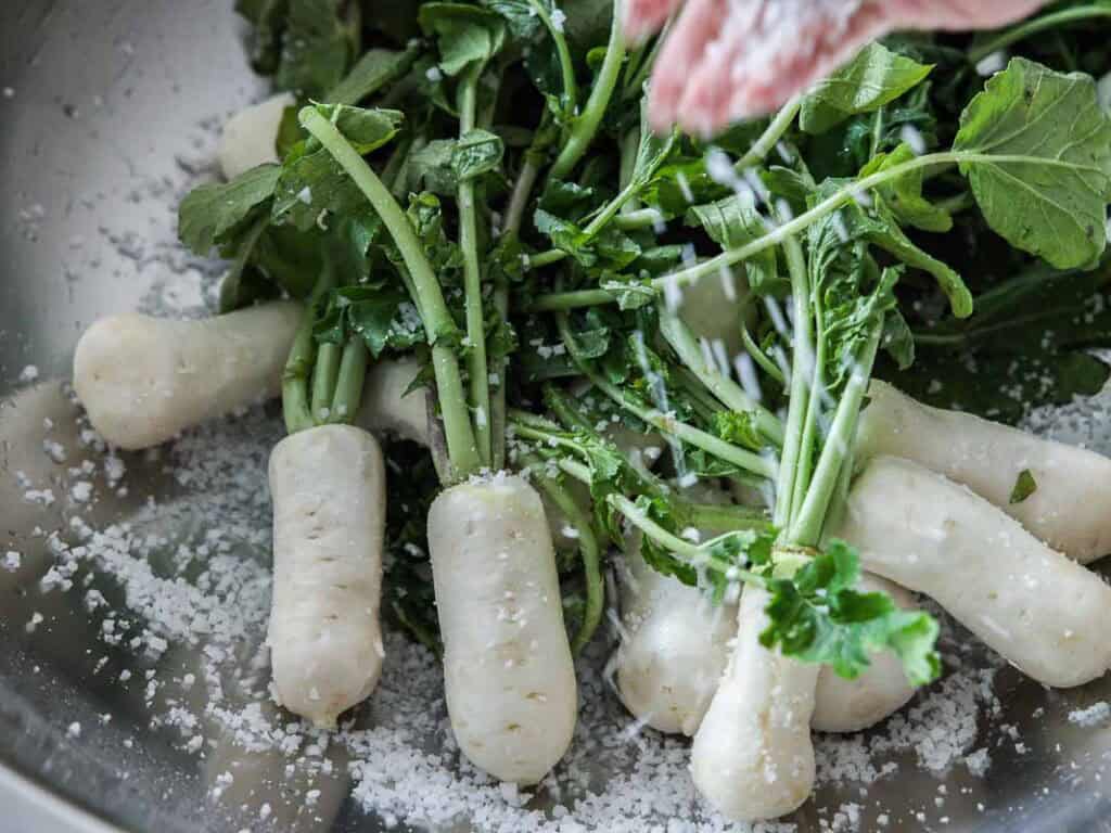 Vegan Ponytail Kimchi aka Chonggak-Kimchi Recipe. 21 Several small white radishes with green leaves are in a metal bowl, being sprinkled with coarse salt from above—the perfect start for preparing Vegan Kimchi or an authentic Chonggak-Kimchi recipe.