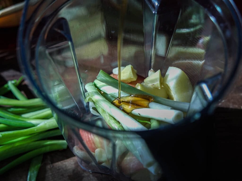 Vegan Ponytail Kimchi aka Chonggak-Kimchi Recipe. 25 Chopped vegetables, including green onions and ginger, in a blender, with a stream of liquid being poured in—perfect for making a Vegan Ponytail Kimchi or Chonggak-Kimchi Recipe. Extra green onions are on the table nearby.