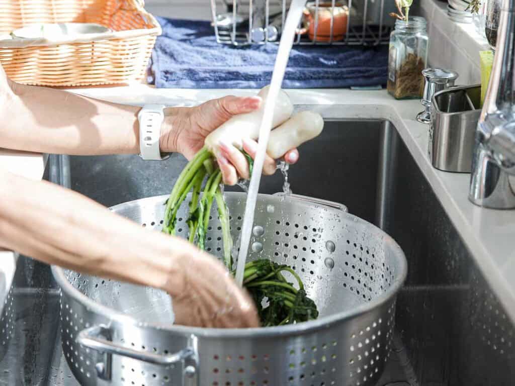 Vegan Ponytail Kimchi aka Chonggak-Kimchi Recipe. 32 A person washes fresh white radishes with green tops in a kitchen sink, prepping them for a Vegan Ponytail Kimchi recipe. Sunlight streams in from a nearby window as the vegetables are held over a metal colander.
