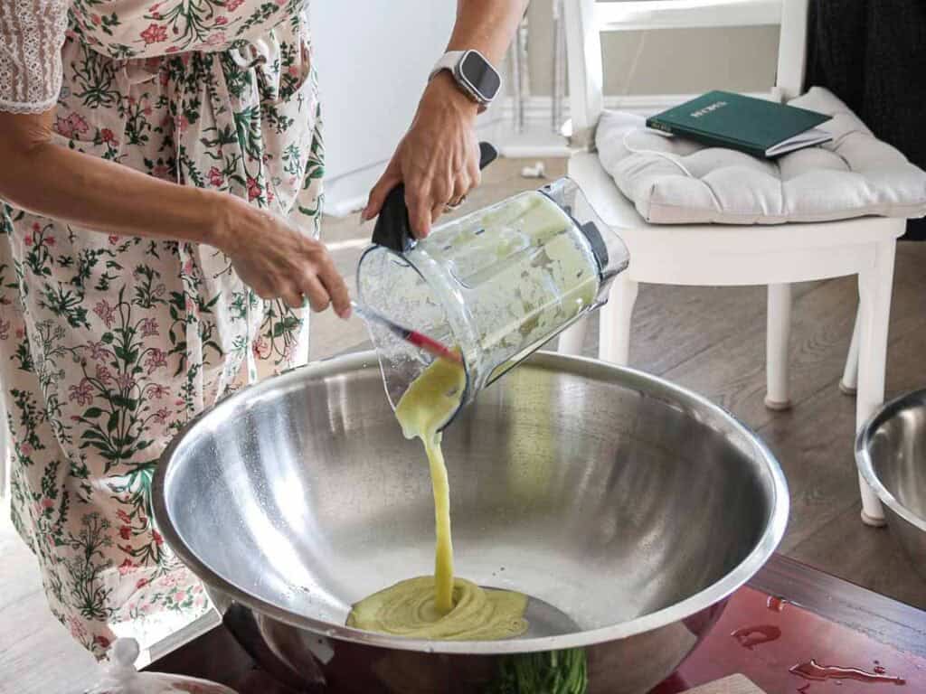 Vegan Ponytail Kimchi aka Chonggak-Kimchi Recipe. 35 A person in a floral dress uses a spatula to pour a thick, yellow-green Vegan Kimchi mixture from a blender into a large stainless steel bowl on a wooden table. A white cushioned chair and a notebook are in the background.