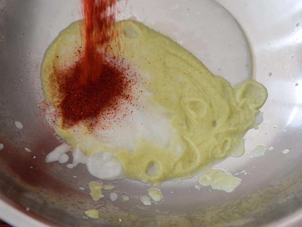 Vegan Ponytail Kimchi aka Chonggak-Kimchi Recipe. 37 A close-up of a metal bowl with melted butter and a pile of red chili powder being added, suggesting the preparation of a spice mixture or sauce for a Vegan Kimchi recipe.