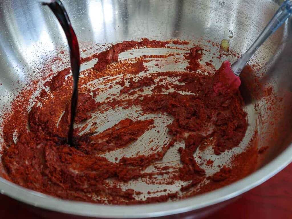 Vegan Ponytail Kimchi aka Chonggak-Kimchi Recipe. 38 A metal mixing bowl with red chili paste, a red spatula, and a dark liquid being poured in—possibly for preparing a Chonggak-Kimchi marinade or sauce as part of a traditional kimchi recipe.