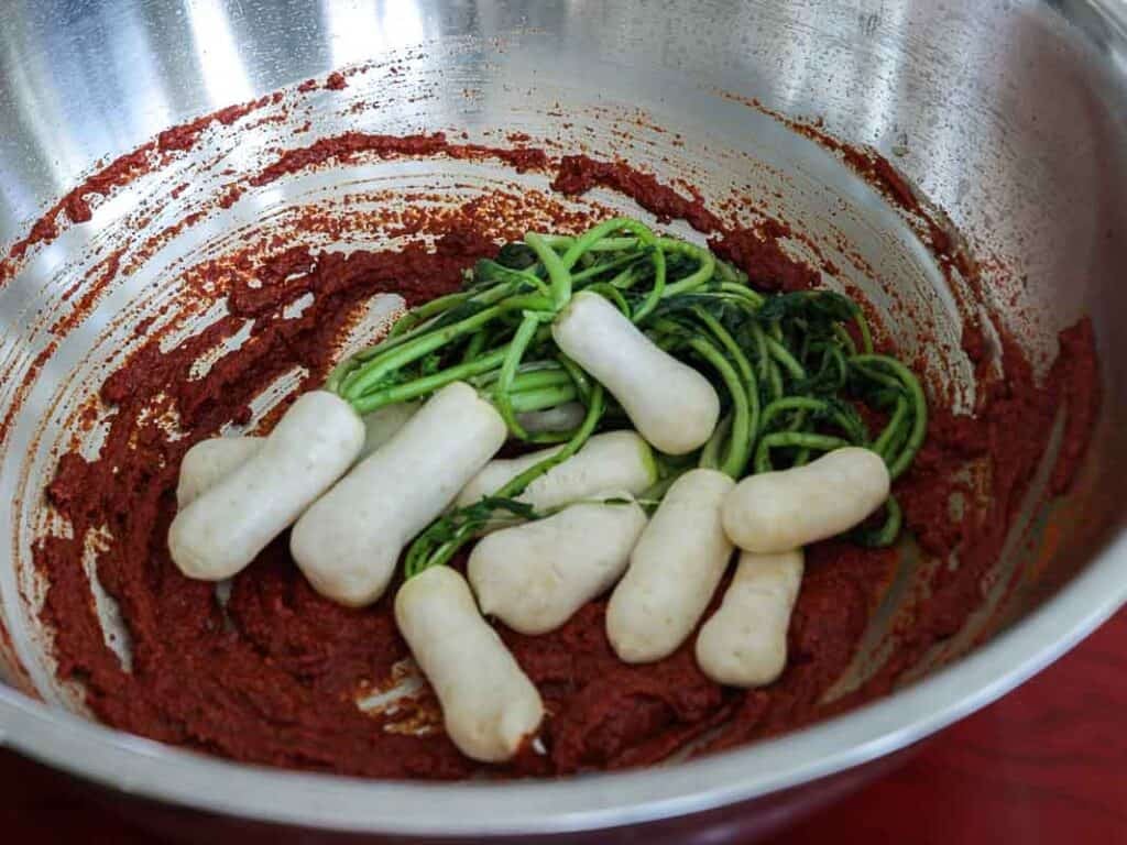 Vegan Ponytail Kimchi aka Chonggak-Kimchi Recipe. 40 A metal bowl containing a red paste, vegan kimchi, several white baby radishes, and green leafy vegetables, partially mixed together.