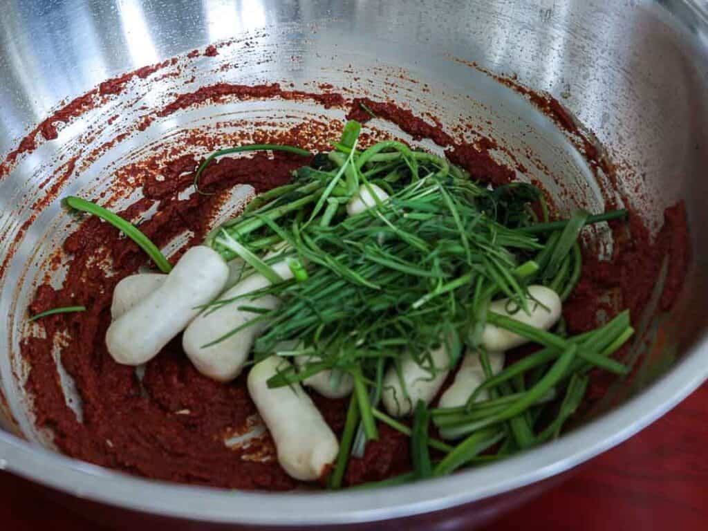 Vegan Ponytail Kimchi aka Chonggak-Kimchi Recipe. 41 A metal mixing bowl contains thick red seasoning paste, baby radishes, and a pile of fresh green chives, ready to be mixed together for a flavorful Vegan Kimchi recipe.