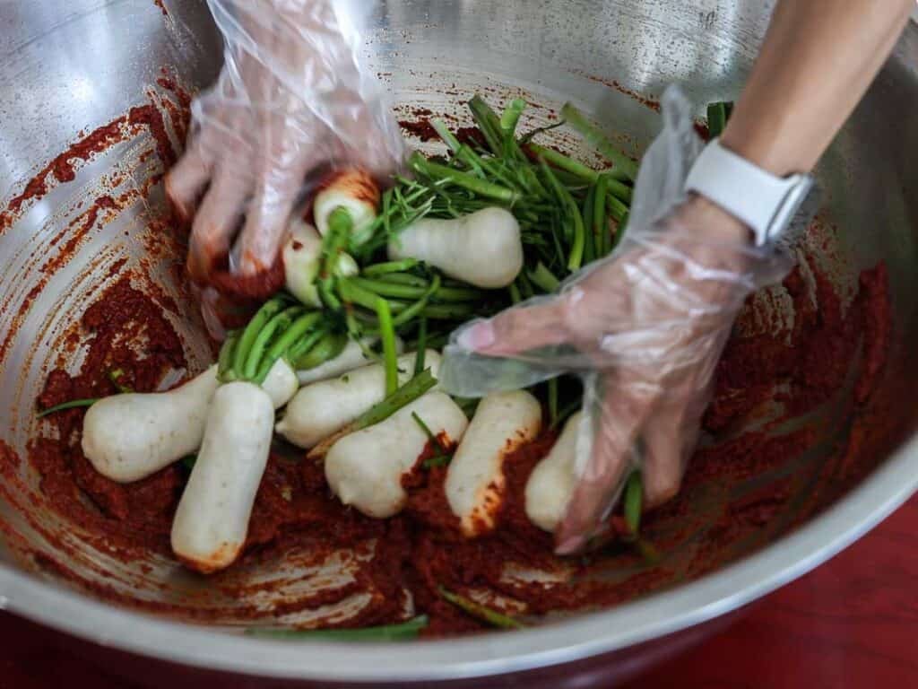 Vegan Ponytail Kimchi aka Chonggak-Kimchi Recipe. 42 Hands wearing plastic gloves mix white radishes and green vegetables with red spicy paste in a large stainless steel bowl, preparing chonggak kimchi. A person wears a white wristwatch on their left wrist.