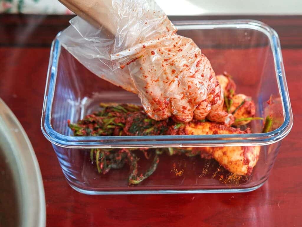 Vegan Ponytail Kimchi aka Chonggak-Kimchi Recipe. 45 A gloved hand mixes vegetables coated with red chili paste in a clear glass container, likely preparing Vegan Ponytail Kimchi, on a wooden surface.