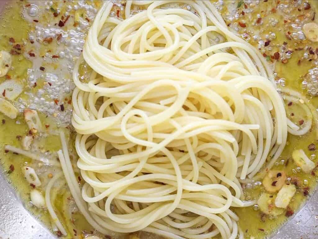 8-ingredient Easy and Delicious Lemon Pepper Pasta. 11 Cooked spaghetti noodles sit atop a bubbling mixture of olive oil, sliced garlic, and red pepper flakes in a pan, preparing for an easy pasta aglio e olio dish that's both simple and delicious.