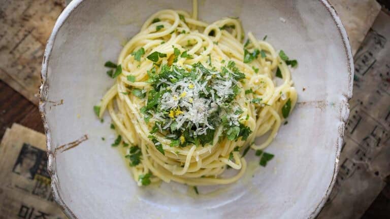 Recipes 8 A bowl of easy pasta topped with grated cheese, chopped parsley, and lemon zest, served in a rustic ceramic dish on a table with old paper sheets underneath.