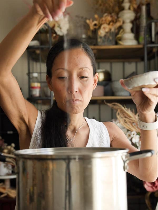 8-ingredient Easy and Delicious Lemon Pepper Pasta. 4 A woman sprinkles salt into a large metal pot in a kitchen, concentrating as she cooks delicious pasta. She holds a small dish in one hand and wears a white sleeveless top and a smartwatch. Shelves with jars and decor are in the background.