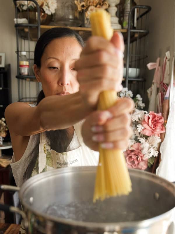 TKV Kollective 4 A woman in an apron holds a bundle of uncooked spaghetti above a pot of boiling water, ready to make an easy pasta recipe in a kitchen decorated with flowers and plants.