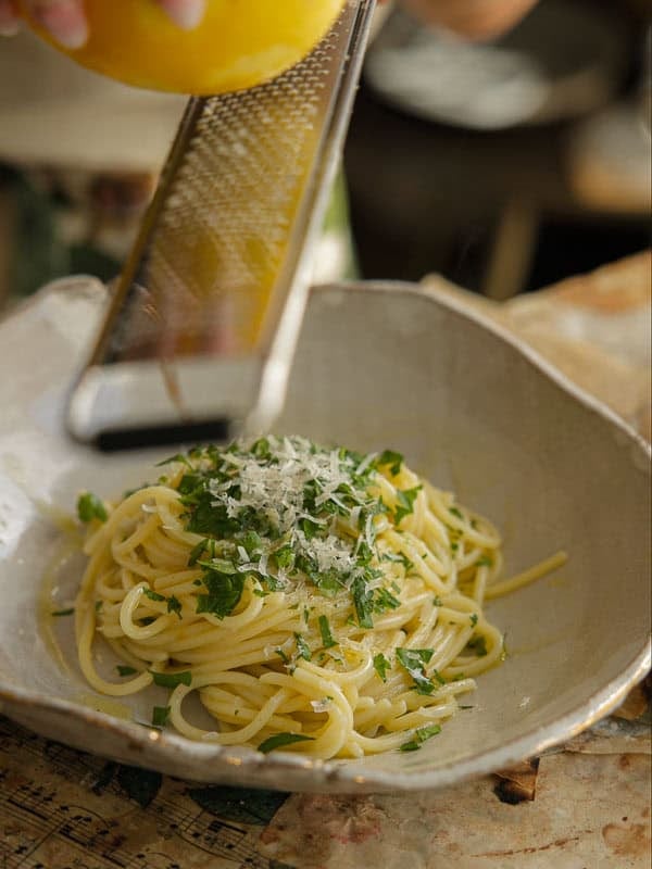 8-ingredient Easy and Delicious Lemon Pepper Pasta. 18 A hand grates fresh lemon zest over a bowl of delicious pasta garnished with chopped parsley and grated cheese. The easy pasta sits in a rustic, shallow bowl on a textured surface.