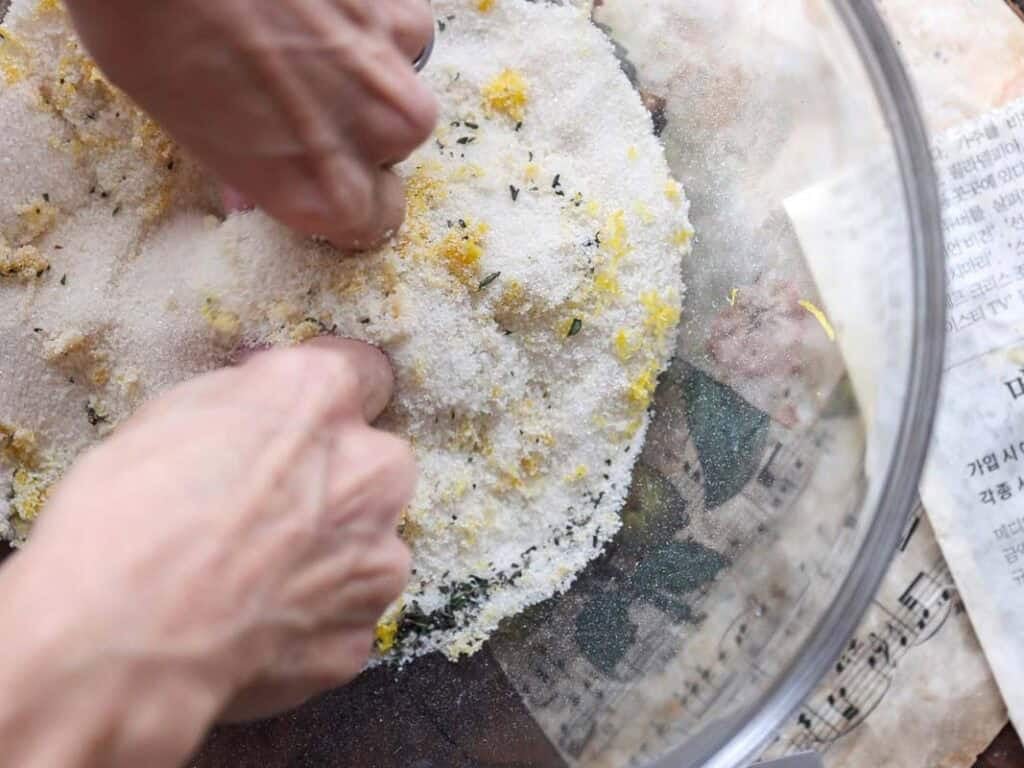 Easy Vegan Lemon Loaf Recipe - Better Than Starbucks 8 Close-up of hands mixing coarse salt, lemon zest, and herbs in a large glass bowl—perfect for topping a vegan lemon loaf. Sheet music and a newspaper peek out from underneath, adding character to the scene.