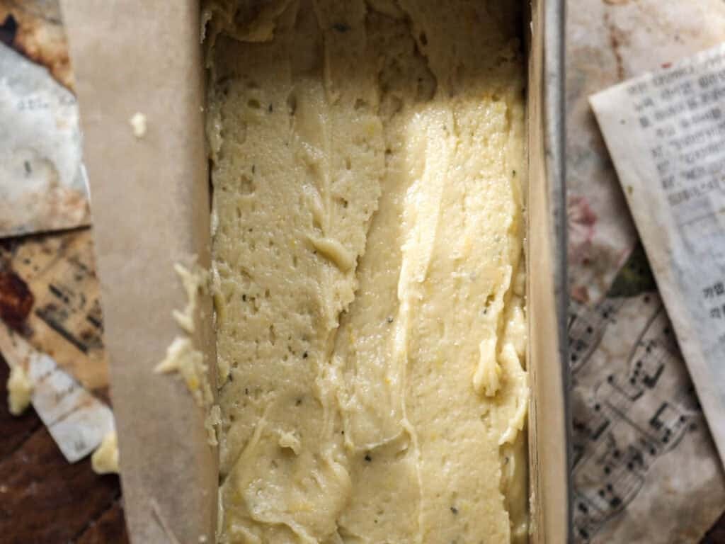Easy Vegan Lemon Loaf Recipe - Better Than Starbucks 17 A close-up of unbaked vegan lemon loaf batter spread evenly in a parchment-lined loaf pan, with vintage sheet music and paper in the blurred background.