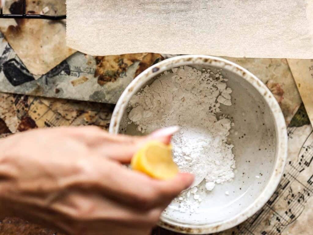 A hand squeezes half a lemon over a bowl filled with white powder, possibly flour or baking soda, prepping ingredients for a vegan lemon loaf on a cluttered, rustic surface scattered with paper and other materials.