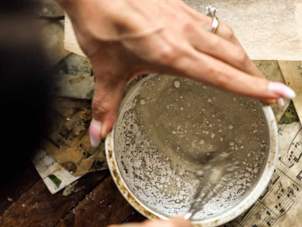 A hand with a ring stirs a white, powdery mixture for a vegan lemon loaf in a bowl using a small whisk, with scattered sheet music and papers visible underneath.