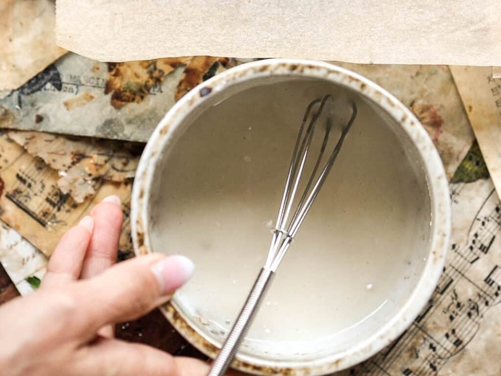A hand holds a ceramic bowl filled with creamy liquid and a small whisk, perhaps prepping a glaze for vegan lemon loaf, resting on vintage, stained sheet music and paper scraps.