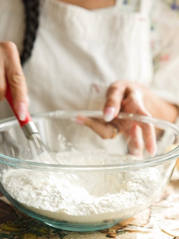 Easy Vegan Lemon Loaf Recipe - Better Than Starbucks 14 A person in a white apron is whisking flour in a glass bowl on a kitchen counter, preparing batter for a vegan lemon loaf. Only their hands and part of their torso are visible.