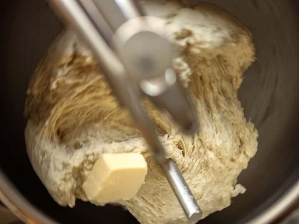 How to Make Viral Salt Bread Recipe (Easy and Vegan). 12 A close-up of bread dough being mixed in a stand mixer bowl, showcasing an easy bread recipe with a metal attachment blending the dough and a chunk of butter on the side—perfect for making viral salt bread at home.