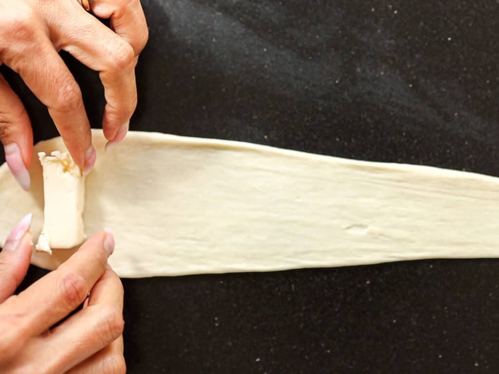 How to Make Viral Salt Bread Recipe (Easy and Vegan). 22 Two hands rolling butter inside a long, thin strip of dough on a dark surface, preparing a pastry or croissant—perfect for trying your favorite easy bread recipe.