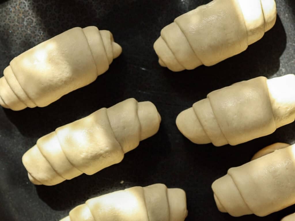 How to Make Viral Salt Bread Recipe (Easy and Vegan). 32 Six unbaked croissant rolls made from dough are placed on a dark surface, with sunlight casting shadows across their smooth, spiral shapes—an inviting scene for anyone seeking an easy bread recipe.