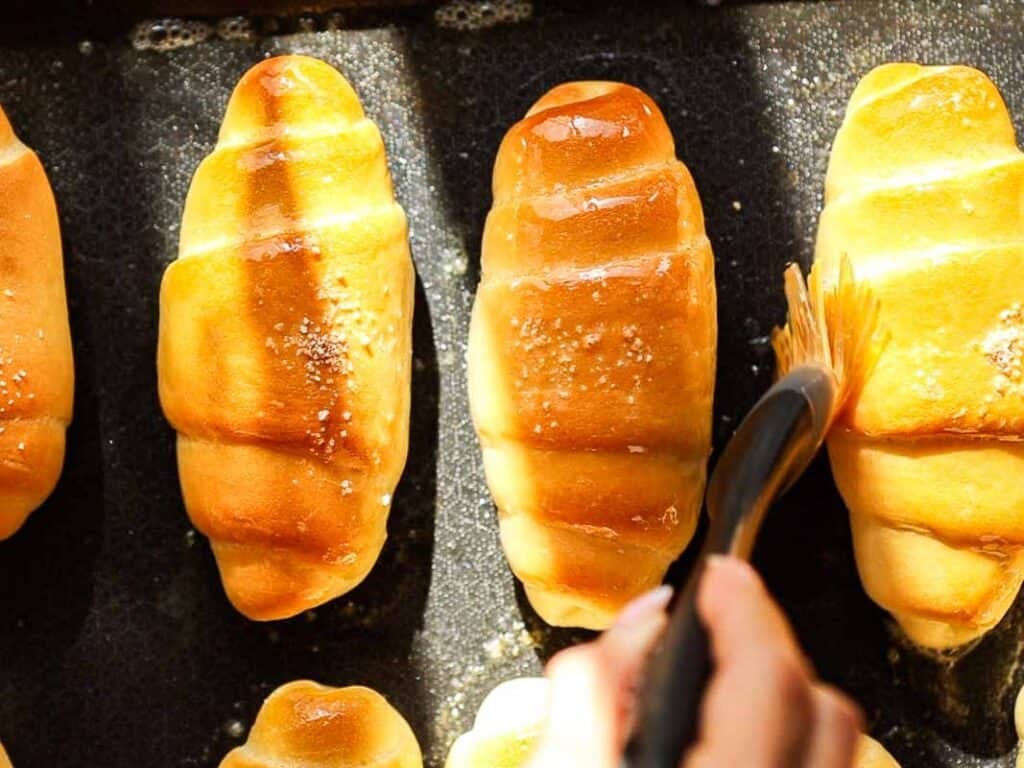 How to Make Viral Salt Bread Recipe (Easy and Vegan). 36 A hand brushes melted butter onto freshly baked, golden brown rolls from an easy bread recipe, giving them a shiny glaze on the baking tray.