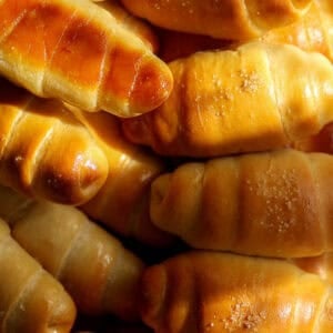 How to Make Viral Salt Bread Recipe (Easy and Vegan). 40 A close-up of several golden-brown, shiny, crescent-shaped bread rolls stacked together, some with a light sprinkling of sugar on top—perfect for trying the latest Viral Salt Bread trend at home.