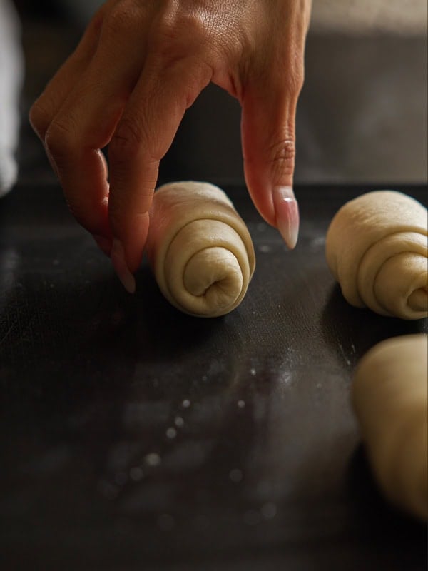 How to Make Viral Salt Bread Recipe (Easy and Vegan). 5 A close-up of a hand with long nails placing a rolled piece of Easy Salt Bread dough onto a dark baking tray with other rolls nearby, ready for baking.