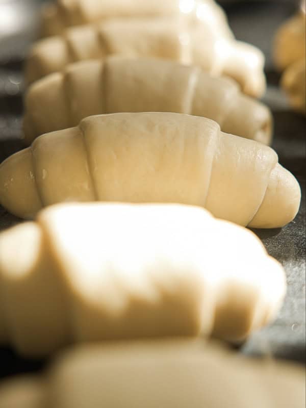 How to Make Viral Salt Bread Recipe (Easy and Vegan). 34 Rows of unbaked croissants made from rolled dough are placed on a baking tray, ready to be baked. This trending viral bread recipe showcases smooth, glossy dough under bright light, promising irresistible fresh pastries.