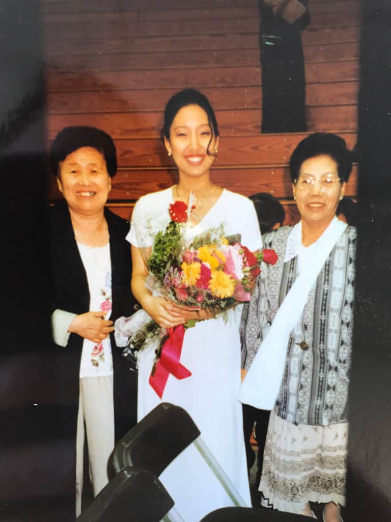 That time I met Barack Obama. 3 Three women stand close together indoors. The woman in the center, dressed in a white gown and holding a bouquet of flowers, is smiling—she once met Barack Obama. The two older women beside her are also smiling. Wooden bleachers are in the background.