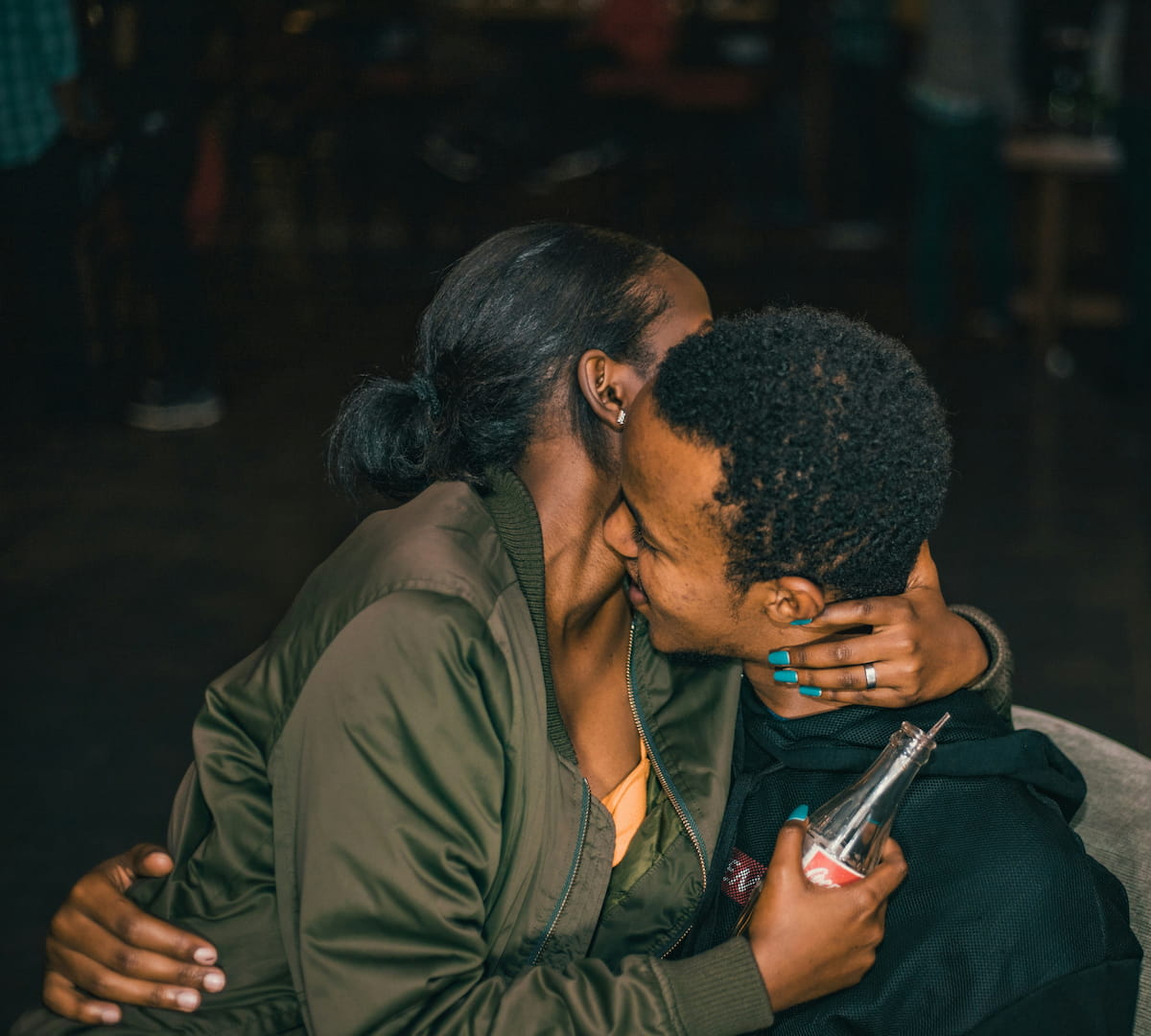 Two people embracing closely. One person in a green jacket whispers to another holding a Coca-Cola bottle. Both have dark hair.